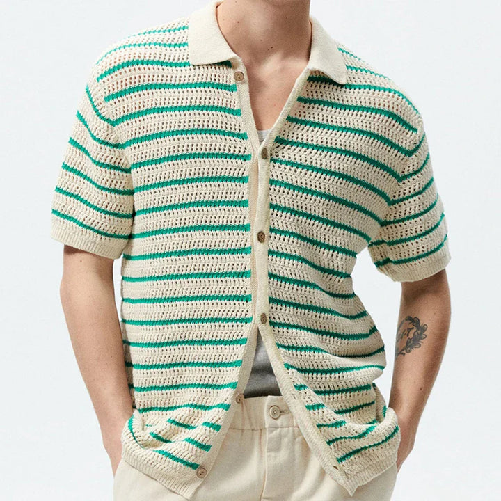 A person wearing a retro knit polo with an open-knit design, featuring green and beige stripes, cream pants, and a tattoo on their left forearm. Their head is not visible, giving the look a fresh streetwear edge.