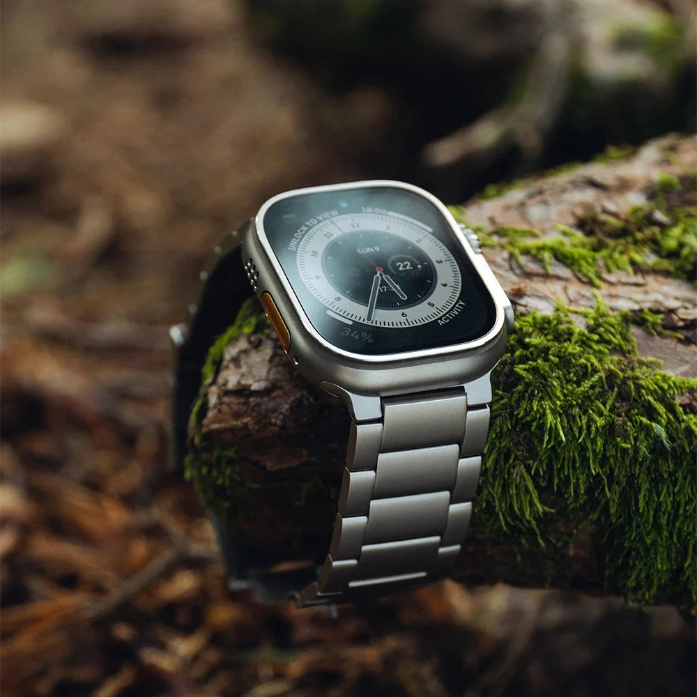 A silver smart watch with an adjustable metal band rests on a moss-covered tree branch in an outdoor, forest-like setting. Crafted from Grade 2 Titanium, it offers lightweight durability while displaying the time and other information.