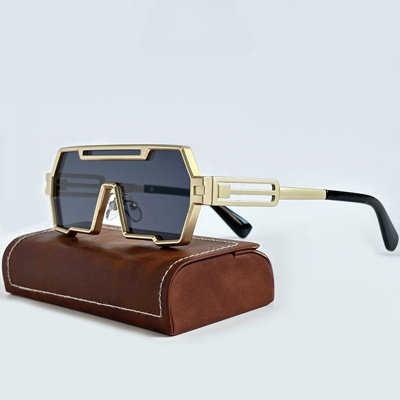 A pair of geometric, gold-framed steampunk sunglasses with dark lenses rests on a brown, rectangular case with white stitching, against a plain light background.