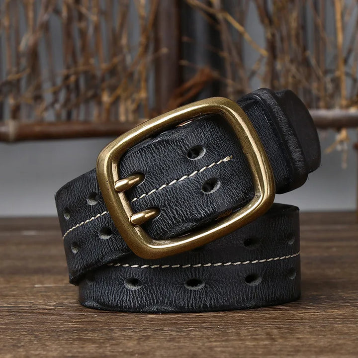 A vintage-inspired belt, this coiled black leather accessory features a gold-tone double prong buckle, white stitching along the edges, and multiple oval holes, displayed on a wooden surface with a rustic background.