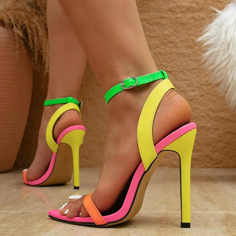 A close-up of a person wearing fashion-forward, high-heeled sandals with vibrant neon colors and an adjustable ankle strap. The stiletto heels stand out against the shiny floor and textured tan background.