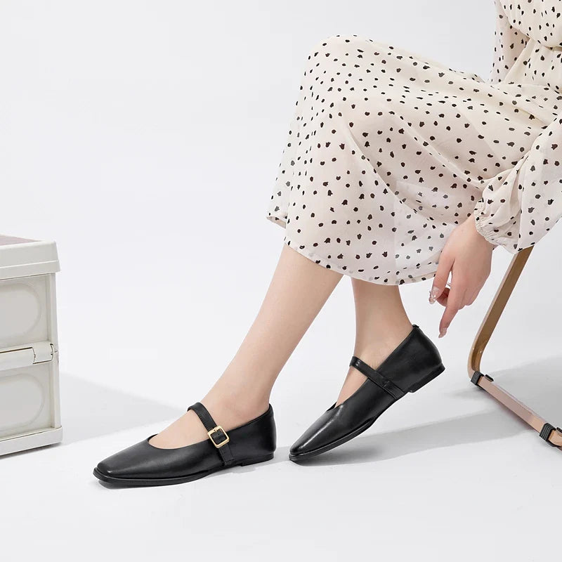 A person wearing a cream-colored, long-sleeve dress with black polka dots sits on a chair, showing off black handmade leather flats with buckled straps. Only the lower half of their body is visible.
