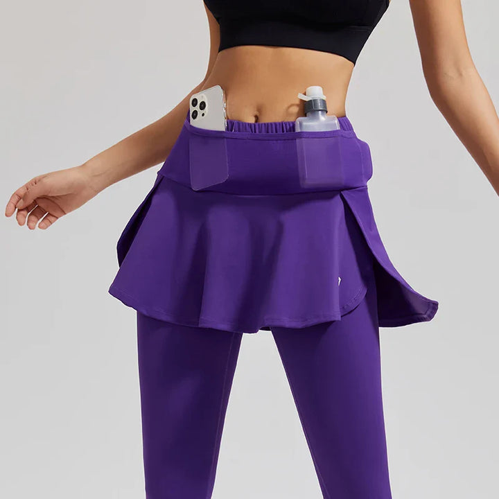 A woman wears high-waisted skirted leggings paired with a black sports bra; the purple sports skirt features stretch storage pockets, holding a smartphone and a bottle.