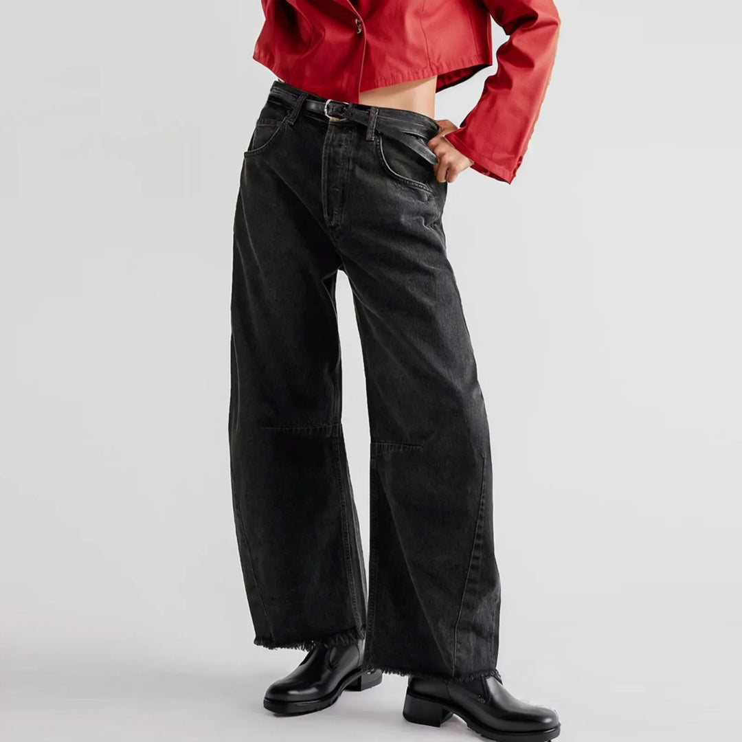 Person wearing black, high waist wide-leg jeans, a red cropped jacket, and black shoes, posing with one hand in their pocket against a plain background.