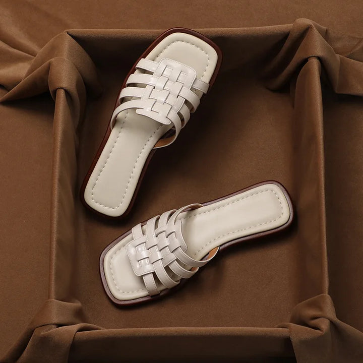 A pair of white, woven Italian leather open-toe slide sandals with square toes displayed on a brown fabric background. These minimalist, modern sandals feature a comfort cushioned insole and are positioned inside a fabric-bordered frame.