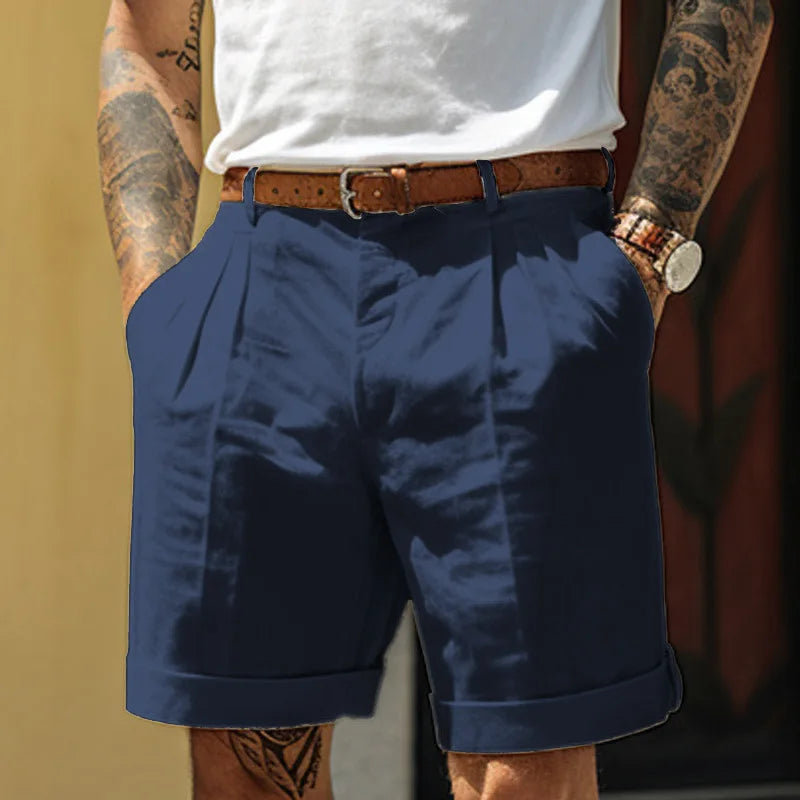 A person wearing a white t-shirt tucked into high-waisted, cuffed navy blue linen shorts with pleats, a brown belt, and a wristwatch. Their tattooed arms and legs show as they keep their hands in the shorts pockets—perfect for summer style.