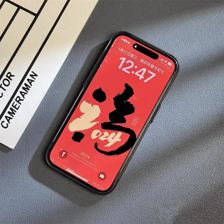 A smartphone with a red wallpaper displaying bold black and gold calligraphy and 2024, protected by a titanium phone case, rests on a gray surface next to a paper labeled DIRECTOR CAMERAMAN. The time on the phone is 12:47.