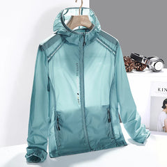A light blue, hooded, zip-up windbreaker jacket made of quick-dry material hangs on a wooden hanger. The jacket has zippered pockets and EXERCISE written vertically on the front. Headphones and pinecones are on the table in the background.
