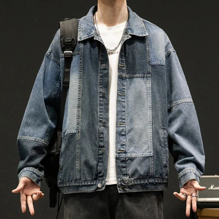 A person wears an oversized fit denim patchwork jacket over a white t-shirt, arms slightly outstretched. With a black backpack on one shoulder, silver rings, and a chain necklace, they stand against a dark background.