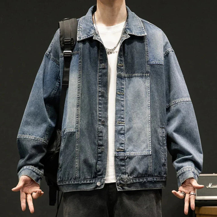 A person wears an oversized fit denim patchwork jacket over a white t-shirt, arms slightly outstretched. With a black backpack on one shoulder, silver rings, and a chain necklace, they stand against a dark background.