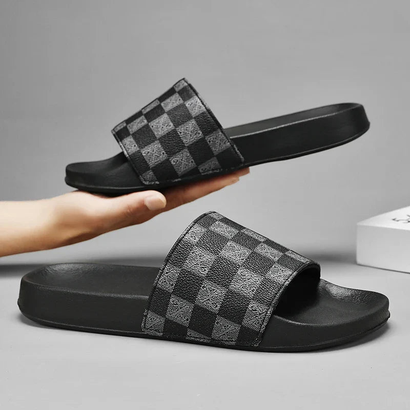 A pair of black checkered sandals with a gray and black checkerboard pattern on the straps, displayed against a plain gray background. One streetwear slide is held up by a hand, while the other rests on a surface.