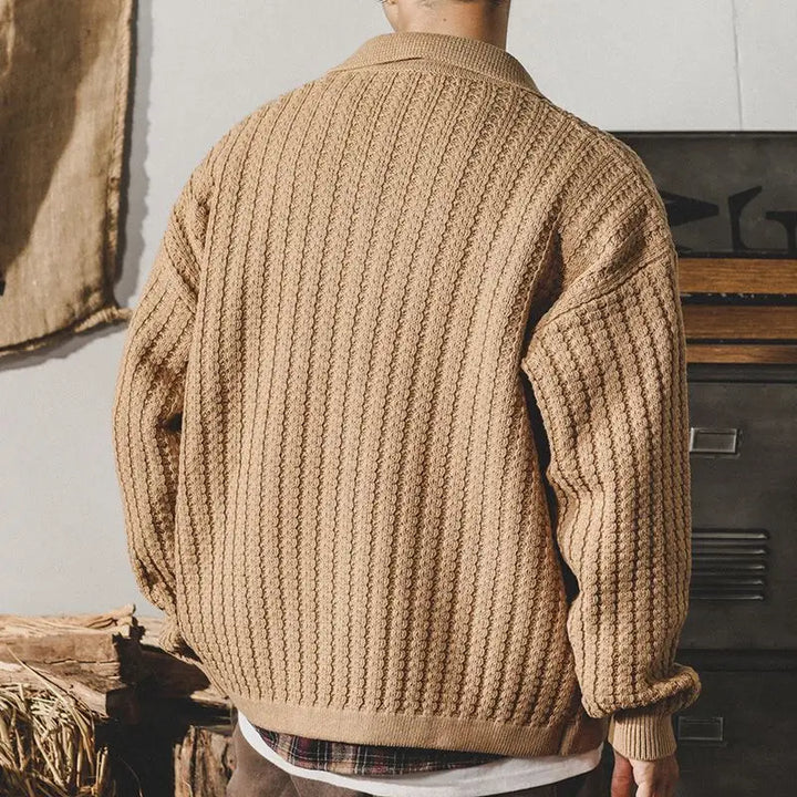 A person stands with their back to the camera, wearing a chunky knit beige sweater. The background features burlap fabric and rustic wooden elements, enhancing the heritage aesthetic.