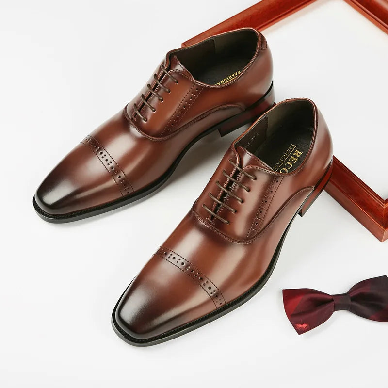 A pair of polished brown Italian leather dress shoes with intricate broguing, placed near a maroon bow tie and a wooden frame on a white surface.