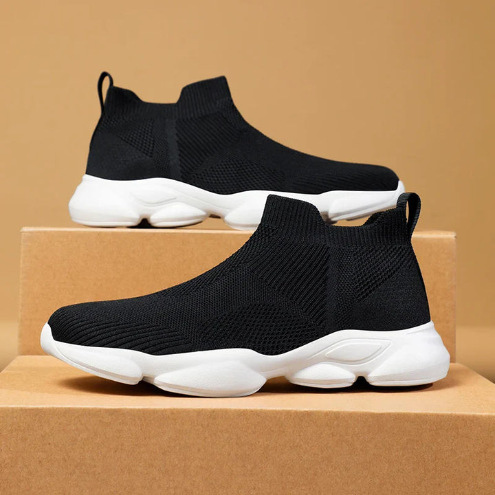 A pair of black slip-on athletic sneakers made from breathable knit fabric with white chunky, non-slip soles displayed on stacked brown cardboard boxes against a beige background.