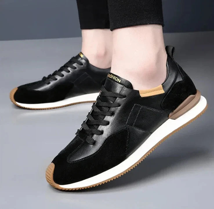 A person wearing black casual sneakers with white soles, tan accents, and FASHION on the tongue, crafted from top-grain leather for durability, stands on a gray surface.