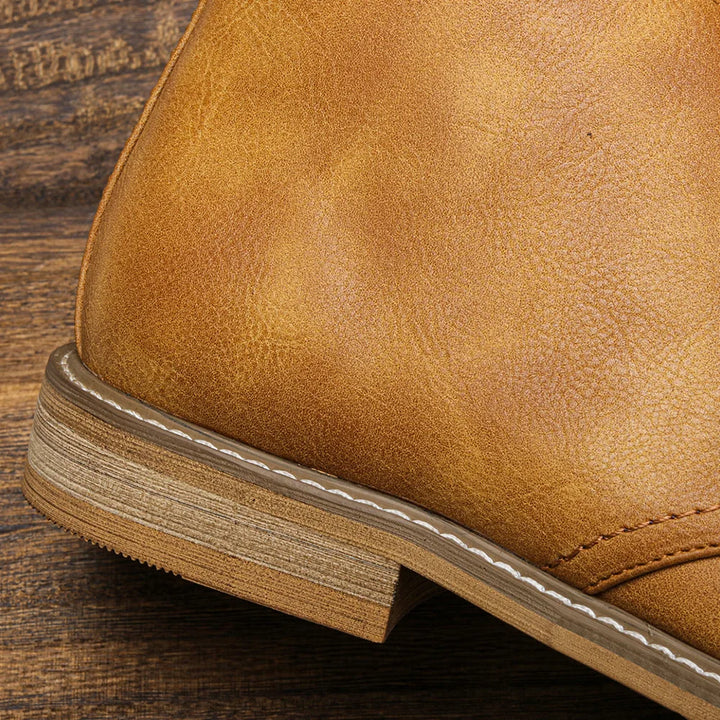 Close-up of the heel and side of a tan leather Western-inspired chukka with visible stitching and a stacked wooden sole, resting on a textured wooden surface.