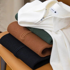 A neatly folded stack of four waffle-knit zip sweaters in wrinkle resistant, stretchy fabric—white, green, brown, and black—rests on a tan chair. The top white sweater features a visible zipper and tag.