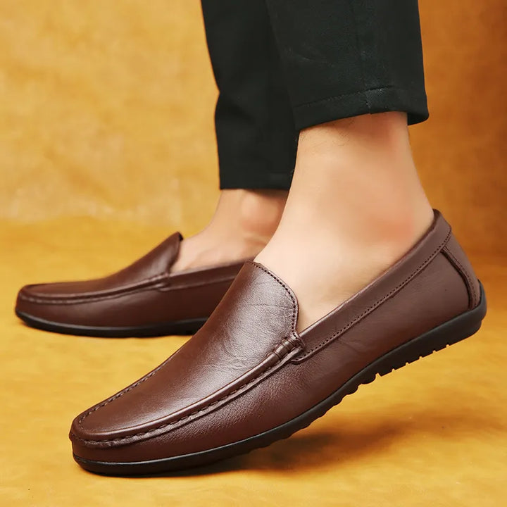 A person wearing luxury shoes with a cushioned footbed and black pants stands on a yellowish floor, showing a side view of the stylish, slip-on loafers.
