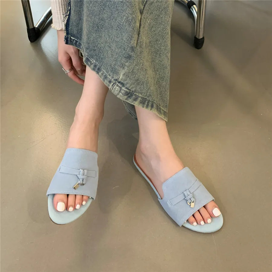 A person wearing light blue open-toe slide sandals with a cushioned insole and wide-leg jeans is seated on a chair, one leg crossed over the other, showing their feet and part of their hand.