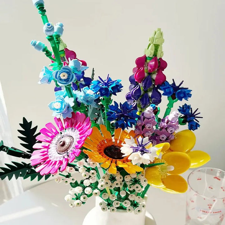 A colorful Wildflower Bouquet of LEGO building blocks, featuring blue, pink, yellow, purple, and white blossoms with green stems and leaves, arranged in a white vase against a bright, neutral background.