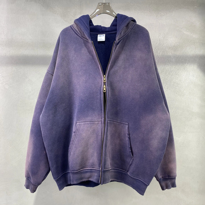 A faded purple zip-up hoodie in a relaxed fit with a double zipper, front pockets, and hood, crafted from thickened fabric and spray-dyed vintage style, hangs on a clear plastic hanger against a light textured wall.