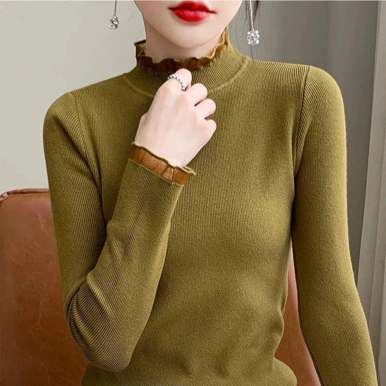 A woman wearing an olive green, long-sleeved, ribbed top with a ruffled mock neck and sleeve cuffs sits on a brown chair. Her hand is raised, showing a ring, and she has red lipstick and dangling earrings.
