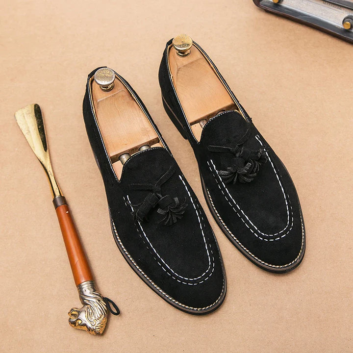 A pair of black suede tassel loafers with white stitching on a tan surface, shown with wooden shoe trees inside and a metal shoehorn with a carved handle beside them, features durable construction for long-lasting wear.