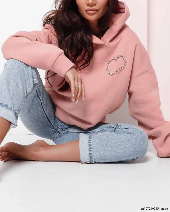 A woman sits cross-legged on the floor, barefoot against a white background, wearing an oversized pink hoodie with a heart design and light blue rolled-up jeans. Her long dark hair is partially visible beneath the hood.