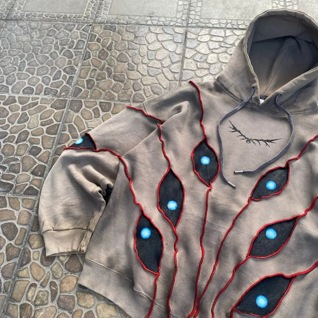 A grey hoodie with a comfortable loose fit features eye pattern patchwork—jagged black patches outlined in red and accented with bright blue “eyes”—laid out on a stone-patterned surface. An eyelash-like graphic decorates the chest area.