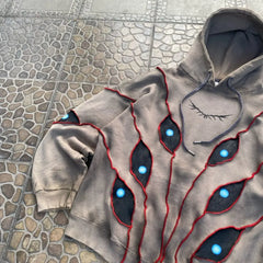 A grey hoodie with a comfortable loose fit features eye pattern patchwork—jagged black patches outlined in red and accented with bright blue “eyes”—laid out on a stone-patterned surface. An eyelash-like graphic decorates the chest area.