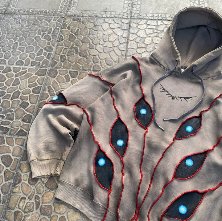 A grey hoodie with a comfortable loose fit features eye pattern patchwork—jagged black patches outlined in red and accented with bright blue “eyes”—laid out on a stone-patterned surface. An eyelash-like graphic decorates the chest area.