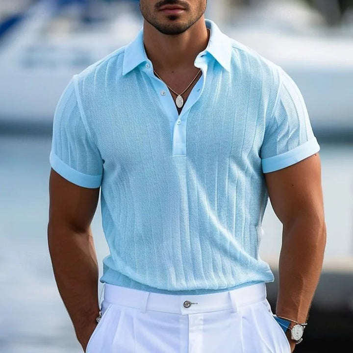 A man in a light blue short-sleeve polo shirt made from lightweight knit fabric and white pants stands outdoors. Sporting a trimmed beard, necklace, and relaxed pose, he appears near a blurred waterfront or marina setting.