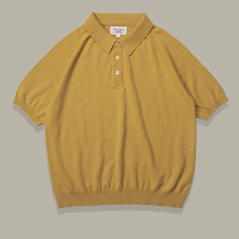 A classic style polo in mustard yellow, this mens polo shirt features short sleeves, a collar, two white buttons, and ribbed cuffs and hem, crafted from soft knit fabric and laid flat on a light beige background.