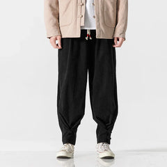 A person wears a beige jacket, white shirt, and black cotton blend sweatpants featuring a drawstring waistband and adjustable fit at the ankles, paired with white sneakers, standing against a plain white background. The persons head is not visible.