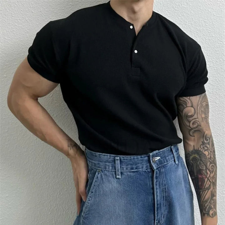 A person stands against a light-colored wall wearing a black, form-fitting shirt with high-waisted blue jeans. One arm is bent at the hip, and the other arm, which has a detailed tattoo, hangs by their side.