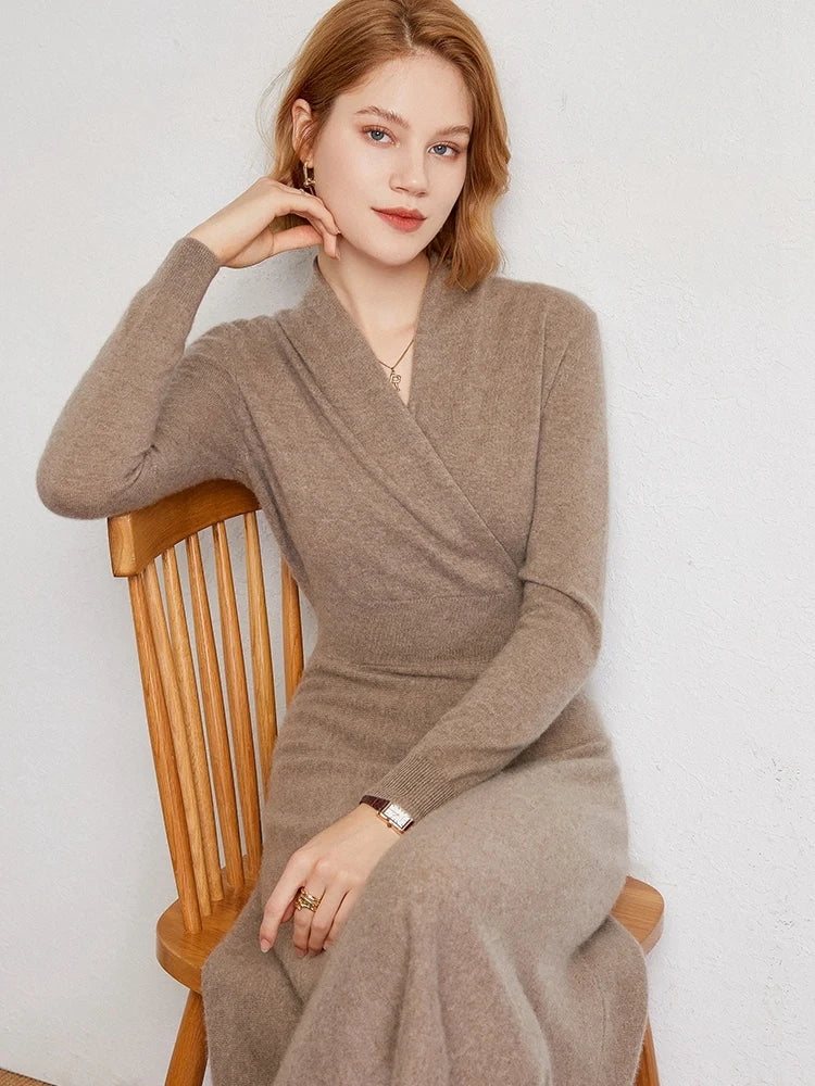 A woman with light brown hair wears a V-neck wrap, beige Matilda Wrap Dress as she sits on a wooden chair, looking at the camera with a slight smile. One arm rests on the chair back and jewelry adorns her wrist and fingers.