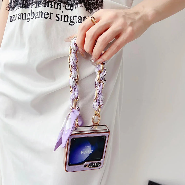A person in a white dress holds a small, square handbag shaped like a retro TV with a purple and gold braided strap. Inspired by the Samsung Galaxy Z Flip, the bag features shockproof protection and a digital clock display on the front.
