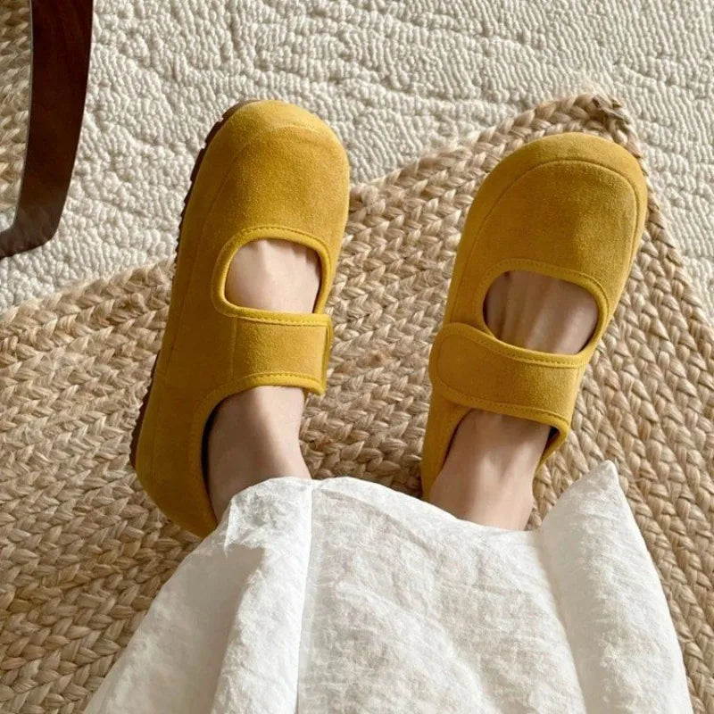 A person wearing mustard yellow Mary Janes rests their feet on a woven mat, with part of a white skirt visible. The textured carpet in the background highlights these stylish women casual shoes.