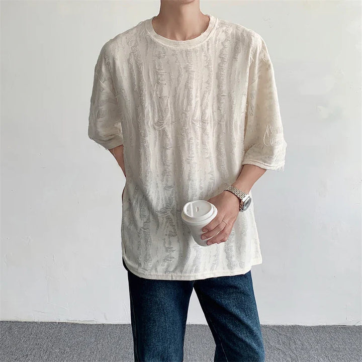 A person in an oversized off-white textured shirt and dark jeans stands against a plain background, holding a white disposable coffee cup—channeling effortless streetwear style.
