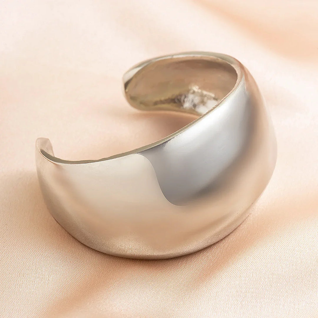 A wide, shiny silver cuff bracelet with a smooth, curved surface and subtle geometric design rests on a soft, light peach fabric background—a striking unisex jewelry piece.
