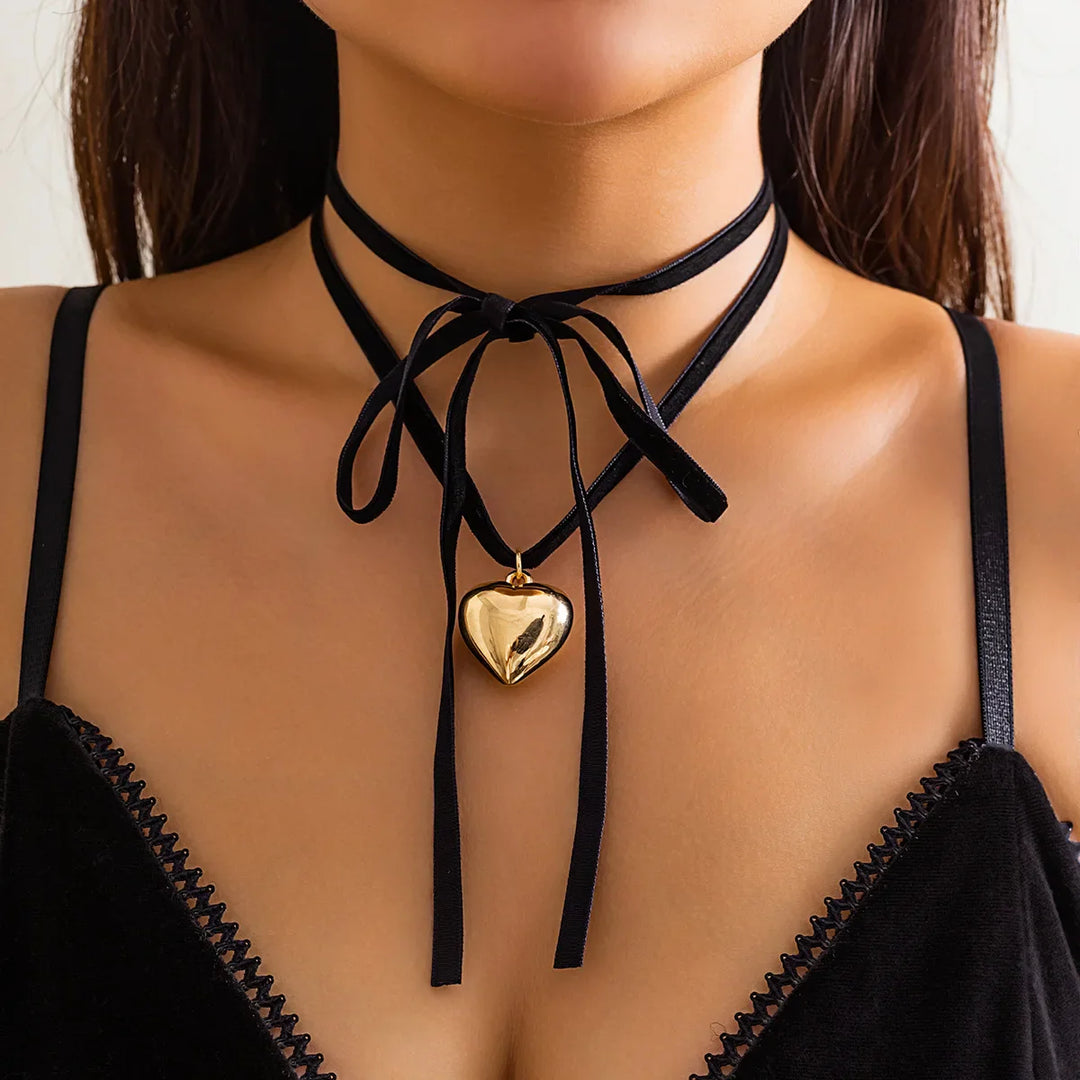 A woman wearing a black velvet strappy top and an adjustable velvet choker tied in a bow, with a shiny gold vintage heart pendant hanging at the center of her neck.