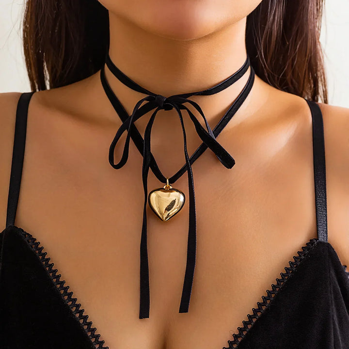 A woman wearing a black velvet strappy top and an adjustable velvet choker tied in a bow, with a shiny gold vintage heart pendant hanging at the center of her neck.