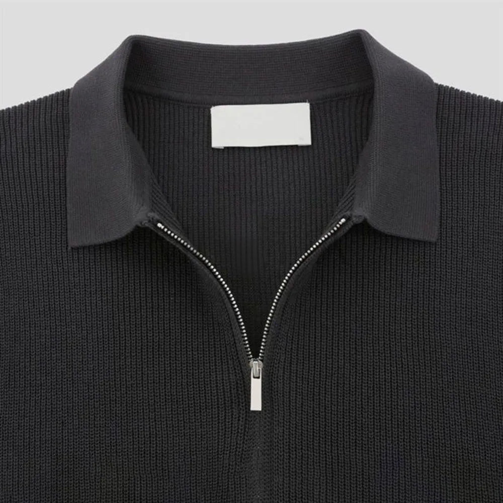 A close-up of a black quarter zip polo with ribbed detailing and a pointed collar, displayed on a plain light background. A blank white label is visible inside the collar.