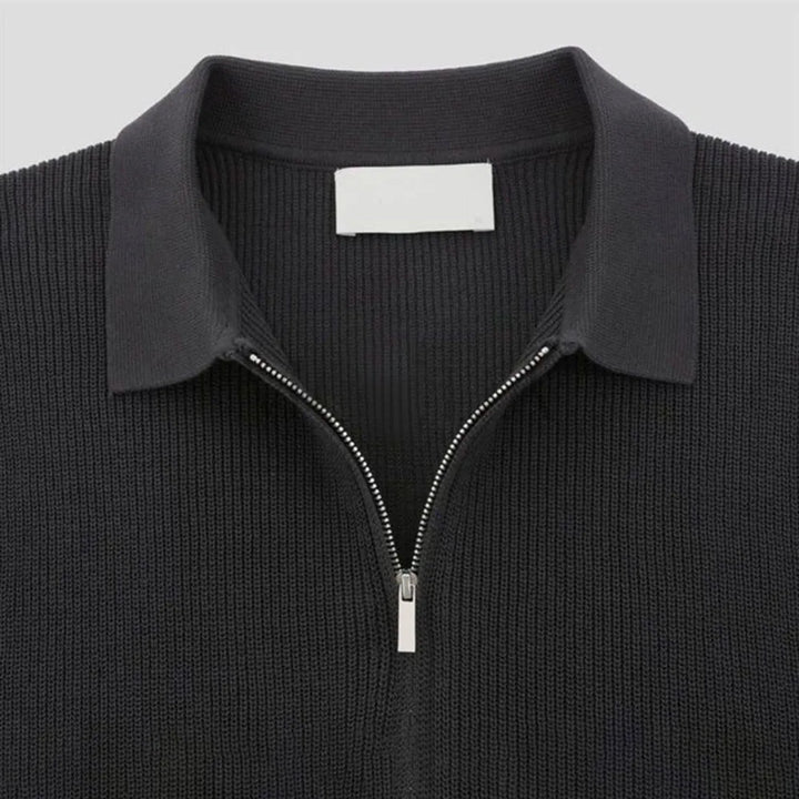 A close-up of a black quarter zip polo with ribbed detailing and a pointed collar, displayed on a plain light background. A blank white label is visible inside the collar.