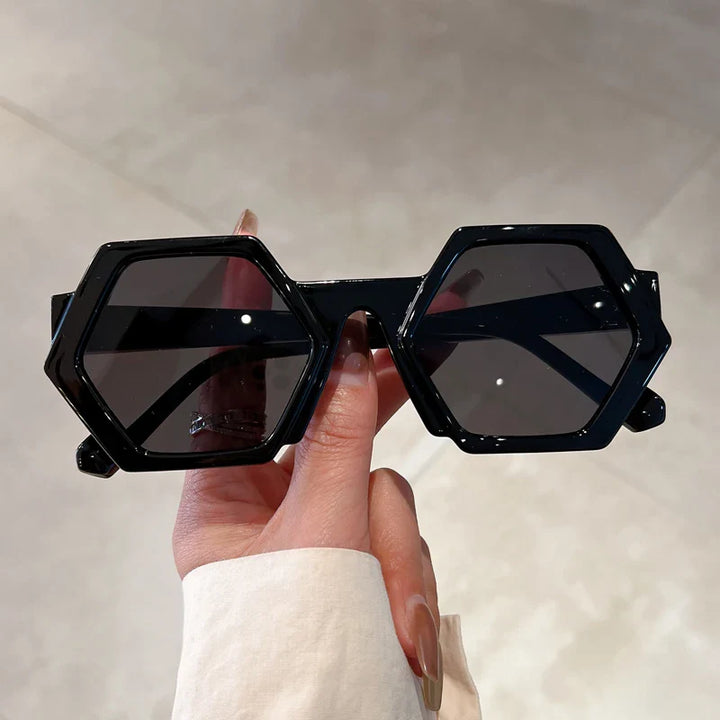 A hand holds trend-forward black sunglasses featuring large, hexagonal frames and dark lenses with UV400 protection against a neutral background.
