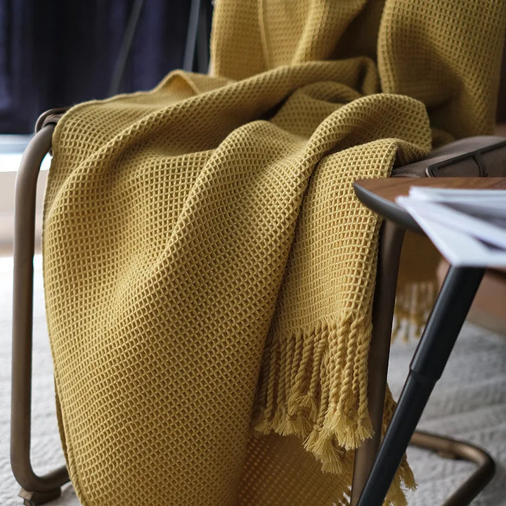 A mustard yellow knit waffle blanket with fringed edges drapes over a brown metal chair, beside a table with scattered papers in a softly lit room, adding a cozy throw blanket boho touch to the decor.
