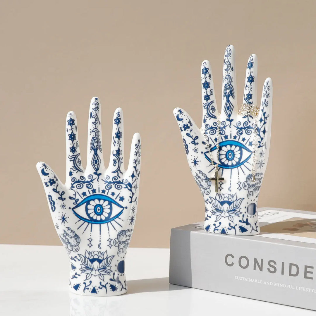 Two ceramic hand sculptures with blue and white intricate designs—eyes, stars, and lotus flowers—serve as unique evil eye decor or a stylish ceramic jewelry stand, displayed upright on a white surface; one rests atop a gray book titled CONSIDER.