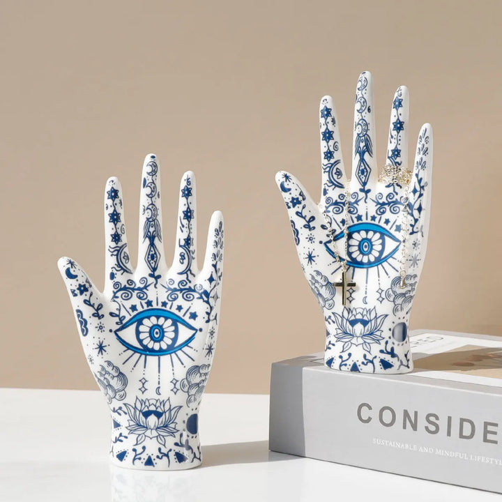 Two ceramic hand sculptures with blue and white intricate designs—eyes, stars, and lotus flowers—serve as unique evil eye decor or a stylish ceramic jewelry stand, displayed upright on a white surface; one rests atop a gray book titled CONSIDER.