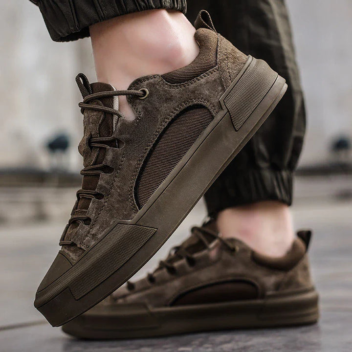 A person wearing brown suede canvas shoes with a cushioned insole and matching brown soles and laces, standing on a paved surface. The individual is dressed in dark, cuffed pants.