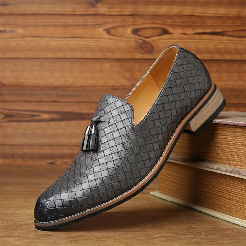 A single luxury footwear black loafer with a basketweave pattern and a decorative tassel, crafted from real leather, displayed on a wooden surface with stacked books against a wooden wall background.